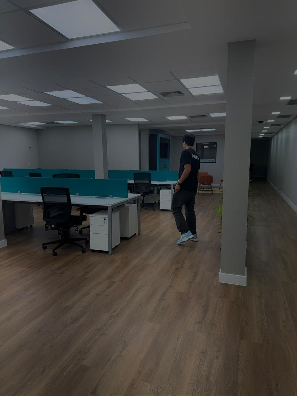 Andar corporativo entregue e em uso, com estações de trabalho, divisórias em azul-petróleo, piso vinílico amadeirado e uma pessoa atravessando o espaço.