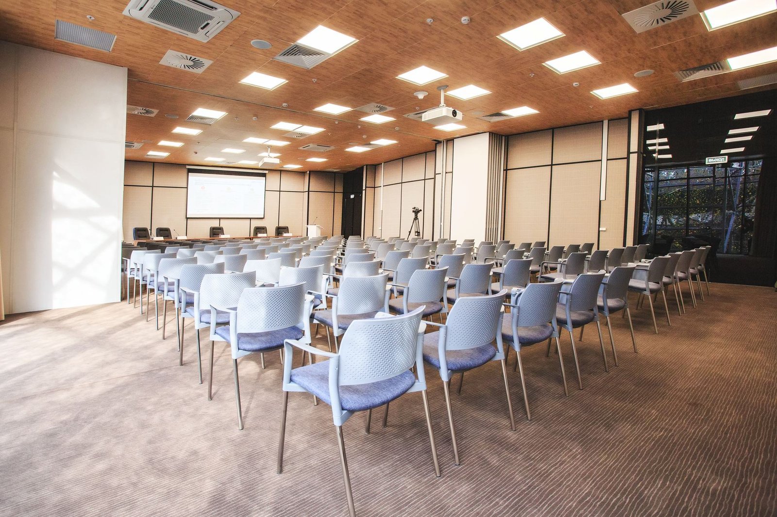 Auditório corporativo montado, com fileiras de cadeiras, tela de projeção e forro de madeira.