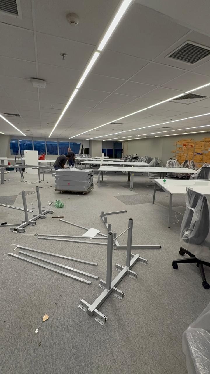Montagem em andamento num andar corporativo, com estruturas de mesa no piso, tampos protegidos e a equipe trabalhando ao fundo.